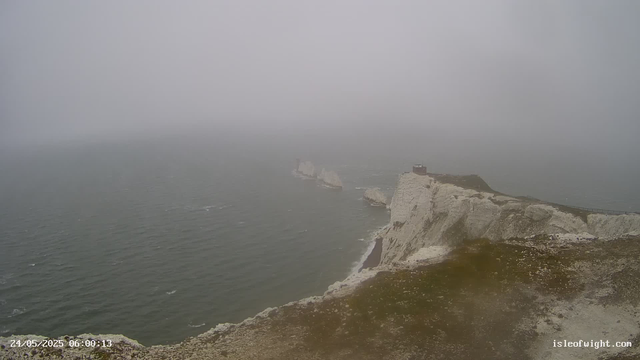 A foggy view of white chalk cliffs along the coastline with a lighthouse at the top. The sea below is calm, and the fog obscures the horizon. There is a pathway along the edge of the cliffs. The image includes a timestamp at the bottom left corner reading "24/05/2025 06:00:13" and a website URL "isleofwight.com" at the bottom right corner.