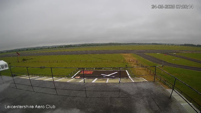 A view of an airport runway and grassy fields on a cloudy day. In the foreground, there is a small helipad with a white helicopter symbol and a red warning sign. The date and time are displayed at the top right corner as "24-05-2025 07:00:14." The bottom left corner shows the text "Leicestershire Aero Club." The overall scene appears to be captured from a webcam overlooking the airfield area.