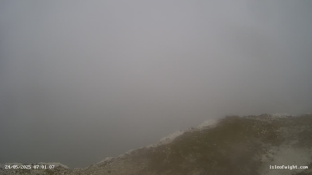 A foggy landscape with the edge of a rocky, muddy surface in the foreground. The background is obscured by thick fog, making it difficult to see beyond the immediate area. The bottom left corner shows a timestamp of "24/05/2025 07:01:07," and the bottom right corner displays "isleofwight.com."