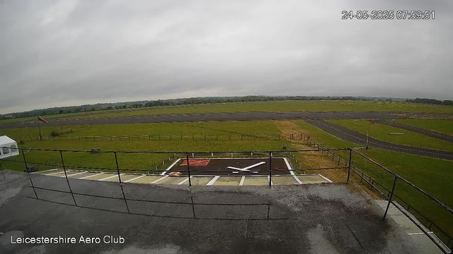 A view from a webcam at Leicesterhire Aero Club shows an overcast sky with gray clouds. In the foreground, there is a small helipad marked with a white "H" on black surface, surrounded by a safety fence. The taxiway and runway are visible beyond the helipad, extending into a flat grassy area with some small wooden fencing. There is a flagpole with a flag on the left side of the image, near a white tent or shelter. The timestamp on the image indicates it was taken on May 24, 2025, at 7:59 AM.