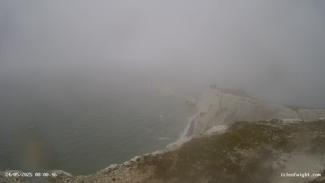 A foggy view of a coastal landscape with white chalk cliffs and a small structure or building on top, partially obscured by the dense fog. The sea is calm with gentle waves, and the foreground shows a rocky, uneven shoreline. The image has a timestamp of 24/05/2025 08:00:46 and a web address "isleofwight.com" in the bottom right corner.