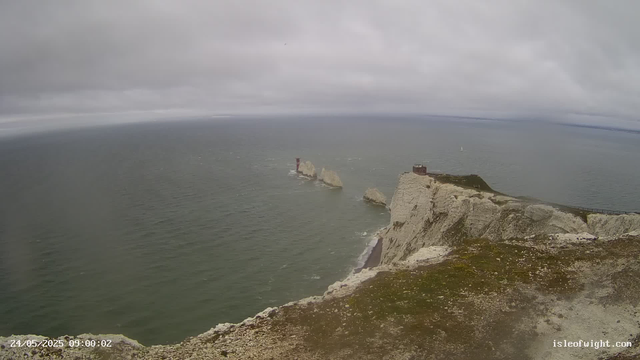 A view of white chalk cliffs and rock formations extending into the sea under a cloudy sky. There is a small building and a pathway along the edge of the cliff on the right side, with three large rocks visible in the water. The scene appears to be taken from a high vantage point, overlooking the coastline and the ocean.