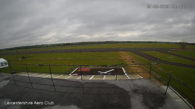 A view from a webcam showing an open grassy field and a runway, with a cloudy sky overhead. In the foreground, there is a balcony or observation deck with a metal railing and a small helicopter landing pad marked with a large white 'H' on a black background. The landing pad is surrounded by white lines. To the left, part of a white vehicle is visible. The image has a timestamp in the top right corner and a label in the bottom left corner reading "Leicestershire Aero Club."