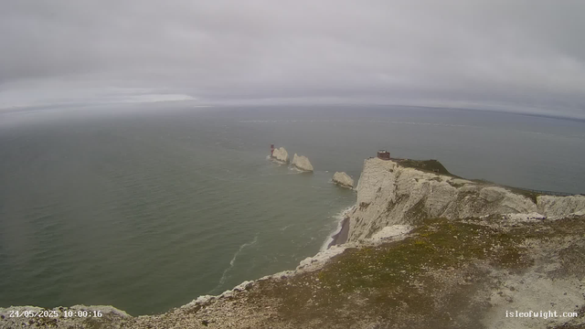 A view of white chalk cliffs with a lighthouse and small building on top, extending into the sea. The sky is overcast with gray clouds, and the sea appears calm with gentle waves. Large rock formations are visible in the water near the base of the cliffs. The image has a timestamp of May 24, 2025, at 10:00:16.