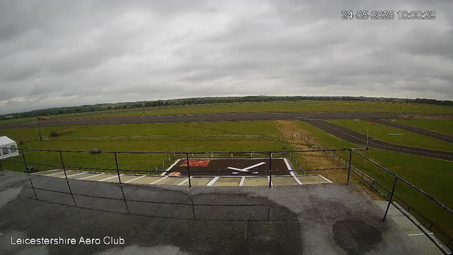 A wide view of an airfield from a webcam, showing a runway with overcast gray skies. In the foreground, there is a helipad with a large white helicopter symbol painted on it, enclosed by a black fence. To the left, part of a small white structure is visible. The surrounding area is grassy, with a few light poles and a distant horizon of fields and trees. The timestamp in the upper right corner reads "24-05-2025 10:00:29," and the bottom left corner displays "Leicestershire Aero Club."