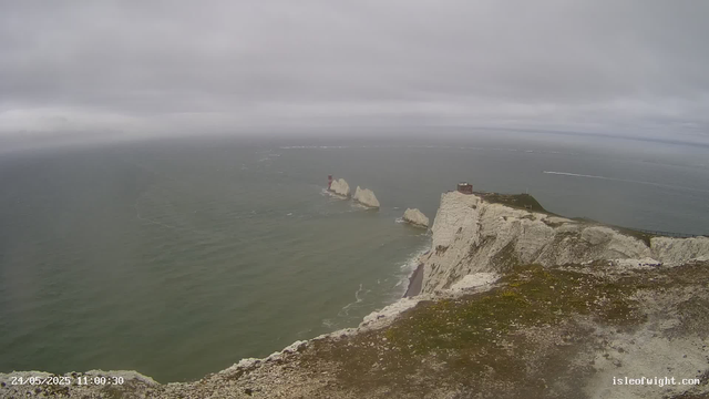 A wide view of white chalk cliffs along the coastline under a cloudy sky. The cliffs extend into the distance, with some rocky outcroppings in the sea below. A small building is visible on top of one of the cliffs on the right side. The ocean appears calm with gentle waves, and a lighthouse stands on a rock formation further out in the water. The photo includes a timestamp at the bottom left corner and a website URL at the bottom right corner.