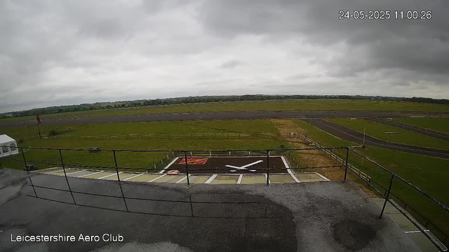 A view from a webcam at Leicestershire Aero Club, showing an airport runway under a cloudy sky. The foreground features a concrete area with a helicopter landing pad marked with a white "H" and a red square, surrounded by a black border. There is a grassy field beyond the landing pad, with a fenced boundary and a small paved taxiway leading towards the runway. The horizon reveals a flat landscape with distant low-lying buildings, and the time and date displayed in the top right corner are "24-05-2025 11:00:26."