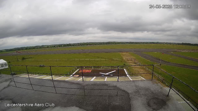 A view from a webcam at Leicesterhire Aero Club shows a cloudy sky over a small airfield with a runway in the distance. In the foreground, there is a rooftop area with a railing, a bike lane, and a small aircraft parked on a marked grid. The landscape is mostly open with green grass, and the weather appears overcast with dark clouds. The timestamp on the image indicates it was taken on May 24, 2025, at 12:00:08.