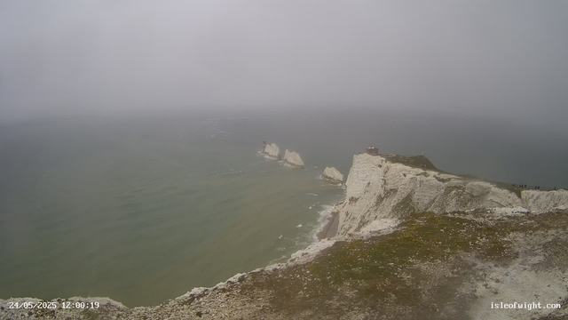 A view of white chalk cliffs along a misty coastline with a few small buildings at the top, extending into the ocean with several large rock formations in the water nearby. The weather appears foggy or overcast.