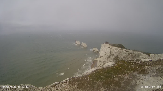 A view of the white chalk cliffs along the coastline, with a small building and a fence on top. The cliffs extend into the distance and meet a hazy, overcast sky over the sea. Several small sailboats are visible on the water in the background. The foreground shows a patch of brown, uneven ground. The timestamp on the image reads "24/05/2025 13:00:42" and the website "isleofwight.com" is displayed in the lower right corner.