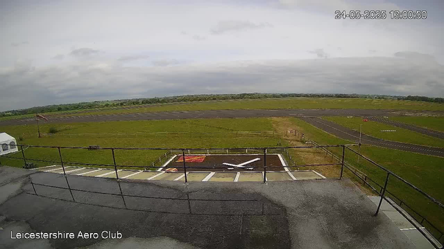 An aerial view of an airfield shows a parking area with a white helicopter landing pad marked with a large white "H." The building has a flat rooftop with a safety railing, and a small aircraft or drone is visible on the ground near the helipad. The field is surrounded by vast open grassy land, with a runway extending into the distance. The sky is mostly cloudy, and the timestamp in the top right corner reads "24-05-2025 13:00:50." The bottom left corner displays the text "Leicestershire Aero Club."