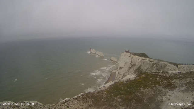 A view of white chalk cliffs along a gray, overcast coastline with a few structures and a pathway on top, overlooking the sea with rough, choppy water under a cloudy sky.