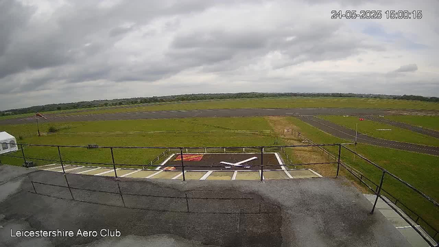 An overcast sky with dark clouds over a flat grassy landscape at Leicestershire Aero Club. A small runway is visible in the distance with a winding taxiway. In the foreground, a viewing platform with a metal railing overlooks the airfield, and there are designated parking spaces or helipad markings on the landing area. The image has a timestamp of 24-05-2025 at 15:00:15.