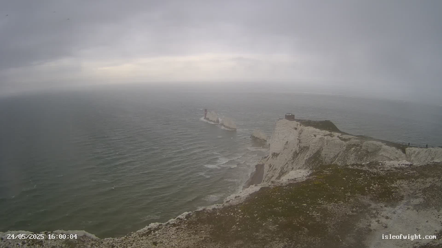 A view of white chalk cliffs extending into the ocean under a cloudy sky. The cliffs have a rugged top with patches of green vegetation, and a small building sits atop the highest point. There is a lighthouse or similar structure further out on a rock formation in the water. The scene appears overcast with gray clouds, and the water below shows gentle ripples. The timestamp in the bottom left corner indicates "24/05/2025 16:00:04," and there is a website URL "isleofwight.com" in the bottom right corner.