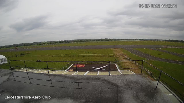 A view from a webcam at Leicestershie Aero Club shows an overcast sky with clouds. In the foreground, there is a small helipad with a white helicopter symbol painted on it and a shaded area with a signage or marking. Beyond the helipad, the tarmac extends into a grassy field with a few small structures, possibly fences or barriers, and an airstrip runs parallel in the background. The timestamp in the top right corner indicates the date and time as 24-05-2025, 16:00:51.