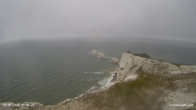 A cloudy, foggy scene of white limestone cliffs along the coastline with the ocean below. Several large sea stacks stand off the shore, and a structure is visible on top of the highest cliff. The water appears calm, with gentle waves lapping against the base of the cliffs. A building with a red chimney can be seen on a smaller outcrop among the sea stacks. The date and time stamp on the bottom left indicate "24/05/2025 17:00:25," and the website "isleofwight.com" is visible on the bottom right.