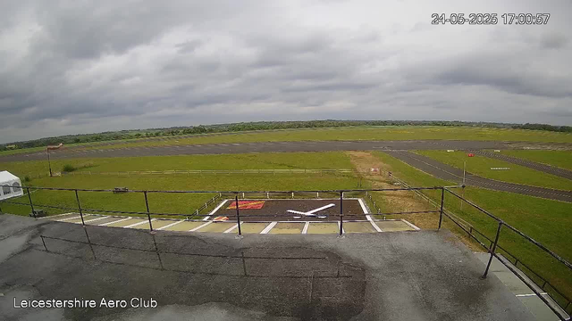 A view from a webcam showing the runway and taxiway of an airfield under cloudy skies. The foreground includes a rooftop with a black railing, and there are designated parking spots for helicopters, with a white helicopter icon painted on the ground. The landscape beyond features green grass fields, a fenced perimeter, and a small glider or aircraft on a distant runway. The timestamp on the image reads "24-05-2025 17:00:57", and the location is identified as "Leicestershire Aero Club."