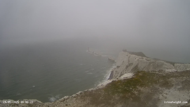 A foggy view of a white chalk cliff coastline with a lighthouse in the distance. The ocean waves crash against the base of the cliffs, and a pathway runs along the top of the cliffs. The overall scene is shrouded in mist, reducing visibility. The timestamp on the image is "24/05/2025 18:00:22" and there is a website link "isleofwight.com" in the bottom right corner.