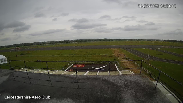 A wide view of an airfield under cloudy skies, with a small helicopter landing pad in the foreground and a runway stretching into the distance. There is a white tent on the left and a flight control building with a railing on the rooftop. The scene appears calm and is part of Leicester Shire Aero Club. The timestamp in the top right corner reads "24-05-2025 18:01:00".