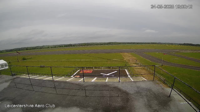 Overcast sky over an empty airfield with a small helicopter landing pad in the foreground, surrounded by a black railing. The runway and grassy areas extend into the distance, with some small signs and markings visible. The image is labeled "Leicestershire Aero Club" in the bottom left corner. The timestamp in the top right corner reads "24-05-2025 19:00:42."