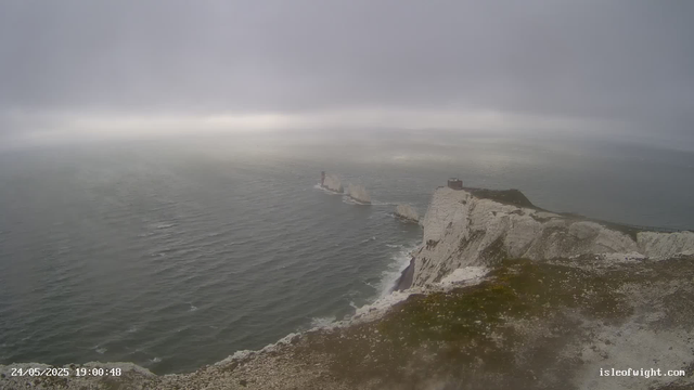 A view of white chalk cliffs overlooking the ocean, with a lighthouse on top of the cliffs and a ship sailing in the distance under a cloudy, overcast sky.