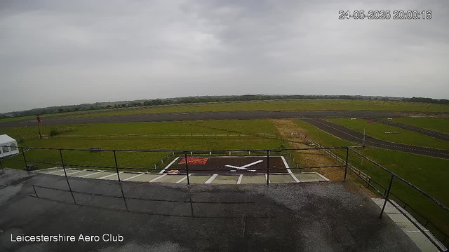 A view of an airfield with a small helicopter on a helipad, surrounded by grassy land and a runway extending into the distance under a cloudy sky. The image has a timestamp in the top right corner and the text "Leicestershire Aero Club" at the bottom.
