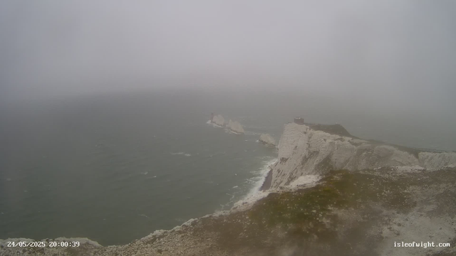 A foggy scene of white chalk cliffs along the coastline extending into the distance over the sea. The sky and the water are gray, with limited visibility due to the fog. A small building or structure is visible on the edge of the cliffs, and a narrow pathway runs along the top of the cliffs. The photo was taken at 20:00 on May 24, 2025, by a webcam on isleofwight.com.