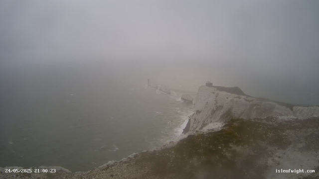A foggy view of white cliffs along a rugged coastline with a lighthouse situated on the edge of the cliffs. The ocean below appears choppy, and the scene is shrouded in mist, obscuring some details in the distance.