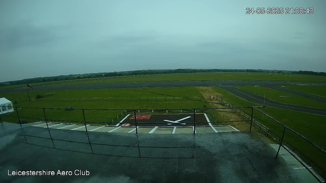 A view from a webcam showing a small aircraft parked on a tarmac at Leicesterhire Aero Club. The aircraft is white with some markings visible on the ground nearby. Surrounding the airfield are green grassy fields, a runway in the distance, and a cloudy sky. The timestamp in the top right corner reads "24-05-2025 21:00:43."