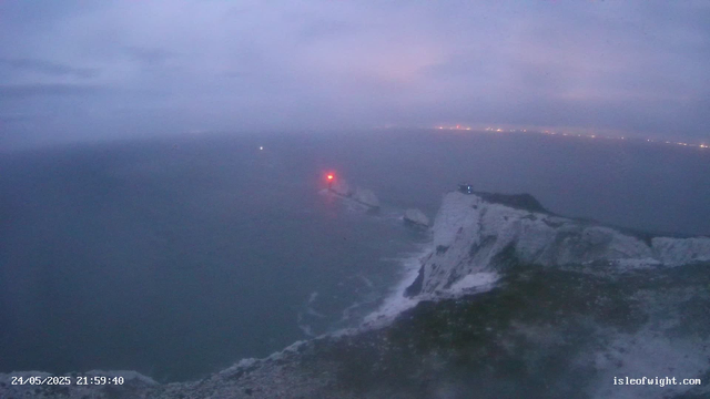 A view from a webcam showing a rocky white cliffside with waves crashing at its base. The ocean extends into the distance under a cloudy, purple-tinged sky at dusk. There are red and white warning lights on the cliff and in the water for navigation. The timestamp reads "24/05/2025 21:59:40" and the website "isleofwight.com" is visible in the bottom right corner.