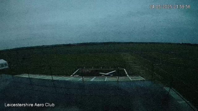 A dark outdoor scene shows the Leicester Aero Club with a large field and a runway visible. There is a small fence around the runway area. The sky appears to be at dusk or dawn, with low lighting and a bluish-gray hue. The time stamp in the upper right corner reads "24-05-2025 21:59:56," indicating evening. The image has a slightly grainy quality.