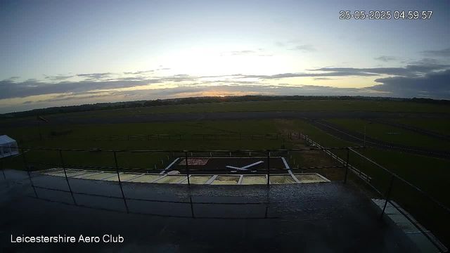 An empty runway at LeicesterShire Aero Club with a green field and cloudy sky in the background during early morning. The date and time are displayed in the top right corner as 25-05-2025 04:59:57.