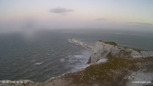 A view of white chalk cliffs with a building and a path on top, overlooking a choppy dark gray ocean. Several large rocks are jutting out of the water in a line extending from the cliff base towards the horizon, with a red lighthouse on one of the rocks. The sky is mostly clear with some scattered clouds, and the lighting suggests early morning or late evening. The image includes a timestamp of 25/05/2025 05:00:15 and a website link "isleofwight.com".
