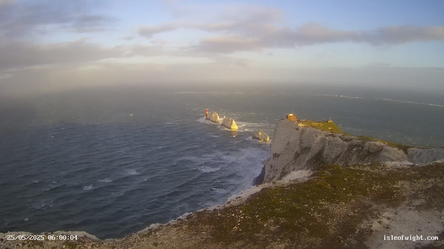 A view of white chalk cliffs with grassy tops extending into the ocean, with a lighthouse visible on a prominent headland. The sea is slightly choppy, and the sky has scattered clouds. The image appears to be taken during the early morning or late afternoon, as the sunlight casts a warm glow on the cliffs. The timestamp on the image reads "25/05/2025 06:00:04," and the website "isleofwight.com" is visible in the bottom right corner.
