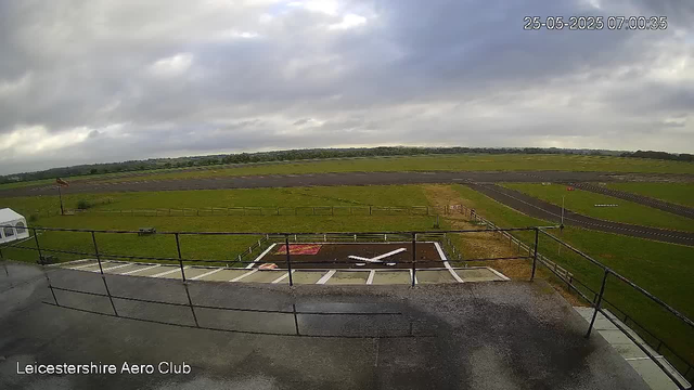 An outdoor view from a webcam showing a grassy airfield with a small helicopter on a landing pad. In the background, there are runways and open fields under a cloudy sky. The timestamp in the top right corner reads "25-05-2025 07:00:35," and the bottom left corner has the text "Leicestershire Aero Club."