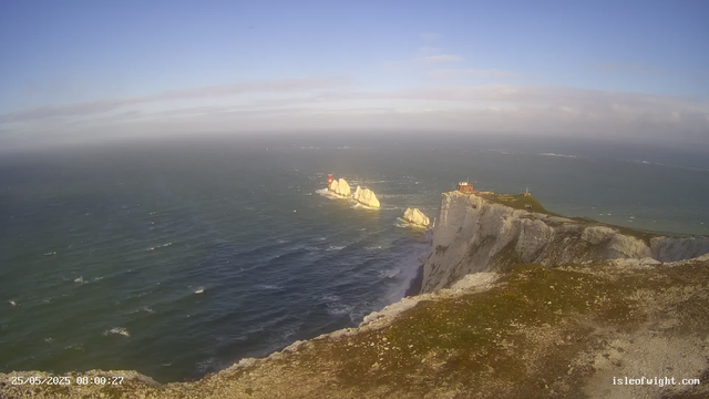 A panoramic view of white chalk cliffs along the coast with a small building and a lighthouse on top, extending into the sea. The ocean has some waves, and there are a few rocks visible in the water near the cliffs. The sky is partly cloudy with patches of blue. The timestamp in the lower left corner reads "25/05/2025 08:00:27," and the website "isleofwight.com" is visible in the lower right corner.