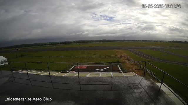A wide view of an airfield showing a runway, grassy areas, and a cloudy sky with some patches of blue. The camera is positioned on a balcony or platform with a metal railing, and the date and time are displayed in the top right corner. The text "Leicestershire Aero Club" is visible in the bottom left corner.