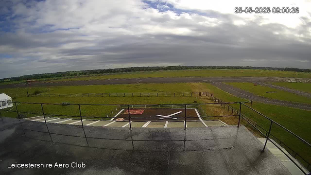 A view from a webcam at LeicesterShire Aero Club showing an outdoor aircraft parking area, a small white helicopter parked on a tarmac helipad, and a grassy airfield with runways in the background under a partly cloudy sky. The timestamp in the upper right corner reads "25-05-2025 09:00:36."