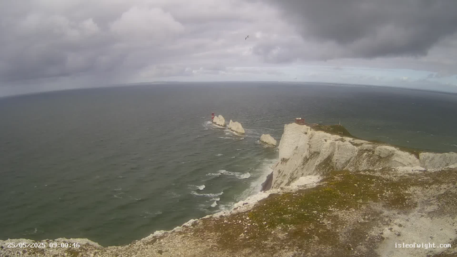 A view of white chalk cliffs extending into the ocean, with a small building or lighthouse on the edge of the cliff. There are large rocks and sea stacks in the water offshore, and a bird is flying above the scene. The sky is overcast with dark clouds. The timestamp reads "25/05/2023 09:00:46," and the website "isleofwight.com" is visible in the bottom right corner.