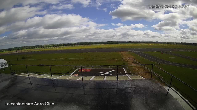A view from a webcam at Leicestershire Aero Club shows a grassy airfield with a helicopter landing pad in the foreground. The sky is partly cloudy with patches of blue, and the landscape in the distance is flat with fields and a few trees. The date and time displayed are 25th May 2025 at 09:59:52.