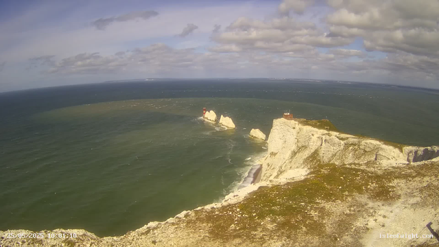 A panoramic view of white chalk cliffs along the coastline under a partly cloudy sky. The ocean is nearby, with some rocky formations and a small lighthouse on a distant rock in the water. A building is situated atop one of the cliffs. The image has a timestamp in the bottom left corner: "25/05/2023 10:01:10."