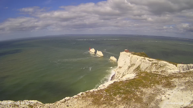 A panoramic view of white chalk cliffs along the coast with a lighthouse and a small building perched on the edge. The ocean waves crash against the rocks below, and boats can be seen in the water nearby. The sky is partly cloudy with patches of sunlight illuminating the scene.
