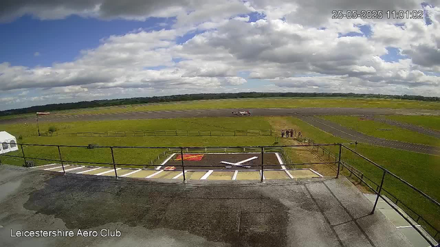 A wide view of an airfield under a partly cloudy sky, with a runway stretching into the distance. In the foreground, there is a helipad marked with an "H" and a small parking area with white lines. A black railing runs along the edge of a platform or balcony from which the photo is taken. In the background, there are grassy fields, a windsock indicating wind direction, and a small fenced area with a few horses. The timestamp in the upper right corner reads "25-05-2025 11:01:02," and the bottom left corner displays "Leicestershire Aero Club."