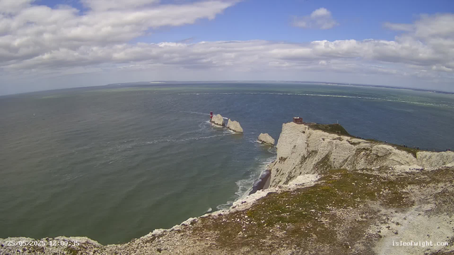 A wide view of white chalk cliffs along the coastline, with a small lighthouse on a rock formation in the sea and a building on the highest point of the cliffs. The ocean extends to the horizon under a partly cloudy sky.