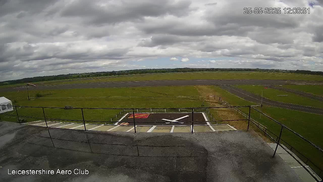 A view from a webcam at Leicestershire Aero Club shows a cloudy sky over a flat grassy airfield with paved runways and taxiways stretching into the distance. In the foreground, there is a helipad marked with a large white "H" and safety markings, along with a small landing pad area. A metal railing surrounds the edge of the platform from which the photo is taken. The timestamp in the upper right corner indicates the date and time as May 25th, 2025, at 12:00:51.