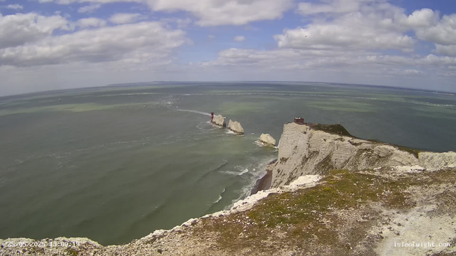 A view from a webcam showing white chalk cliffs extending into the sea, with a lighthouse on one of the smaller rock formations and a building on the main cliff. The ocean is calm with gentle waves, and the sky is partly cloudy with patches of blue. The timestamp in the bottom left corner indicates the date and time as May 25, 2023, at 13:30.