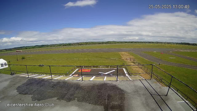 A wide-angle view of a small airfield on a partly cloudy day. In the foreground, there is a black asphalt platform with a railing, and a white aircraft silhouette painted on the ground. To the left, a white tent is visible. Beyond the platform, there is a grassy area with a small fence and a flagpole with a red and white windsock. The runway stretches across the landscape with some additional markings visible. In the distance, the sky is partly cloudy with patches of blue. The image has a timestamp in the upper right corner showing "25-05-2025 13:00:49," and a label at the bottom left reading "Leicestershire Aero Club."