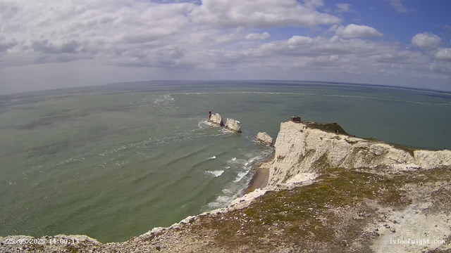 A view from a webcam showing white chalk cliffs beside the sea under a partly cloudy sky. The cliffs extend into the water, with a small structure on top of the cliffs and a lighthouse on a rock formation offshore. Waves are gently crashing against the base of the cliffs, and the horizon stretches far into the distance.