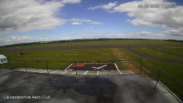 A view from a webcam showing an open grassy field and a runway with a small helipad marked with an "H" in white and red paint. The sky is partly cloudy with patches of blue showing through. In the foreground, there is a concrete area with a metal railing and a few parking spaces. The timestamp in the top right corner reads "25-05-2025 14:00:29," and text at the bottom left says "Leicestershire Aero Club."