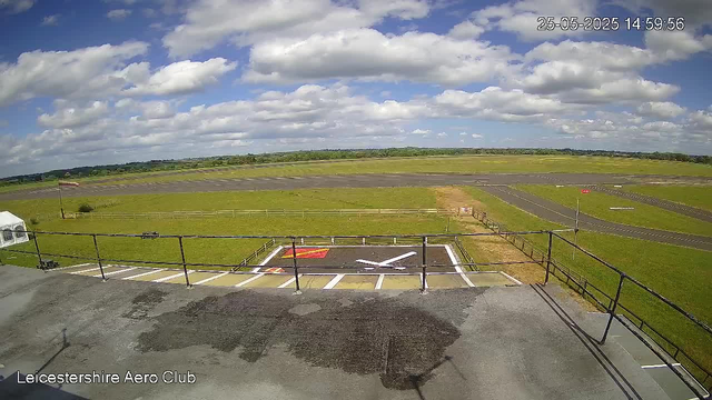A view from a webcam at Leicestershire Aero Club showing a grassy airfield with runways extending into the distance. The sky is partly cloudy with scattered white clouds and a blue sky background. The timestamp in the top right corner indicates the date and time as 25-05-2025, 14:59:56. The foreground includes a small parking area with a designated helipad painted with a white "H" and red markings, surrounded by a black safety barrier. The landscape in the background is flat with some trees and low hills on the horizon.